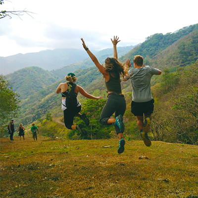 Three students in Costa Rica run and jump across a hilly, forested landscape.